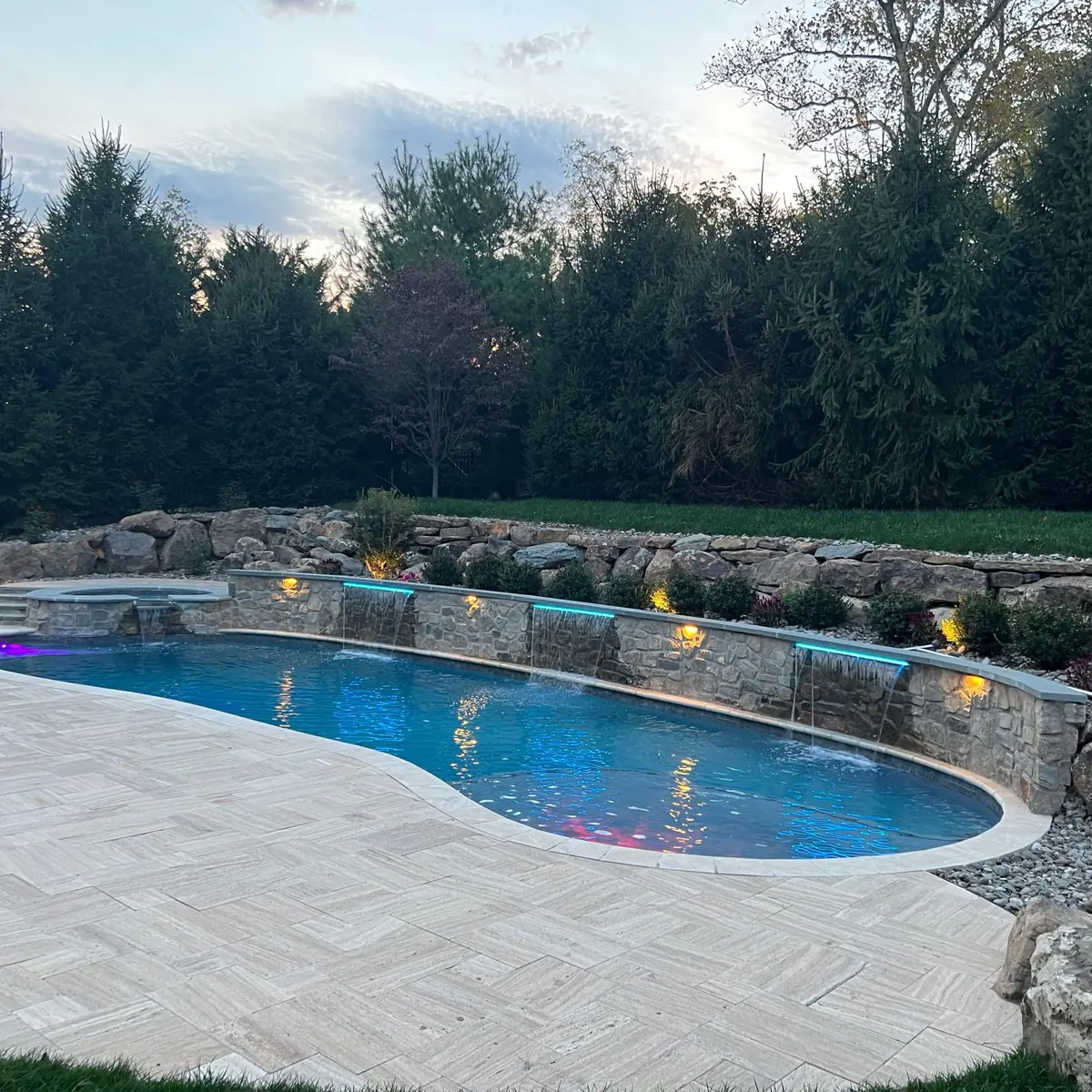Freeform Chester County pool with an illuminated stone waterfall wall at dusk